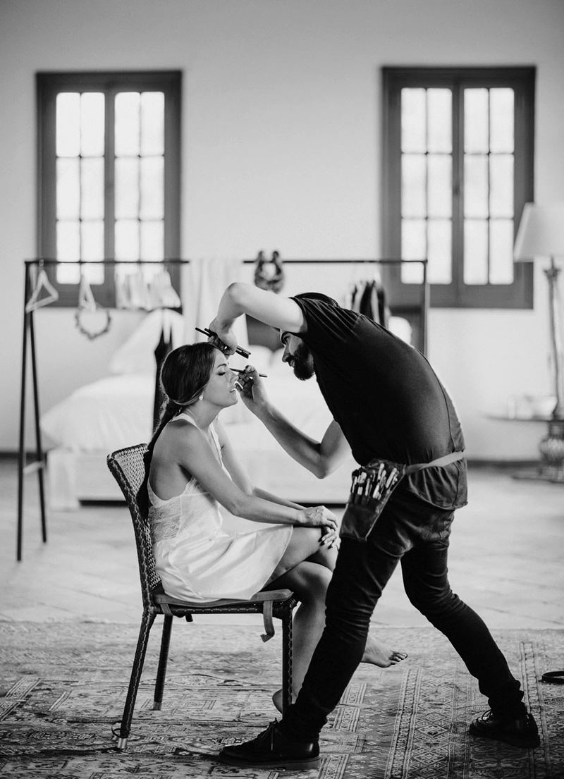 Fotografía de retrato en la que aparecen un maquillador y la novia en la sesión de maquillaje el día de la boda.